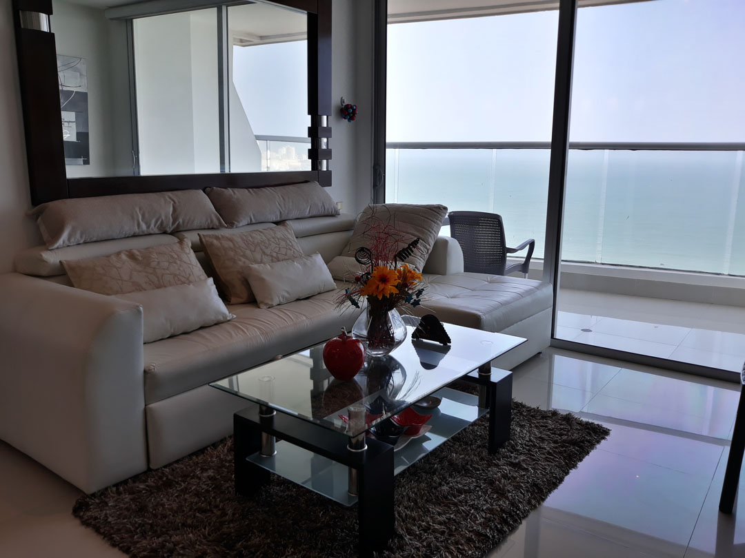sala de estar con sofá de cuero blanco en L y mesa de café, un ventanal da salida a un balcón con vista al mar
