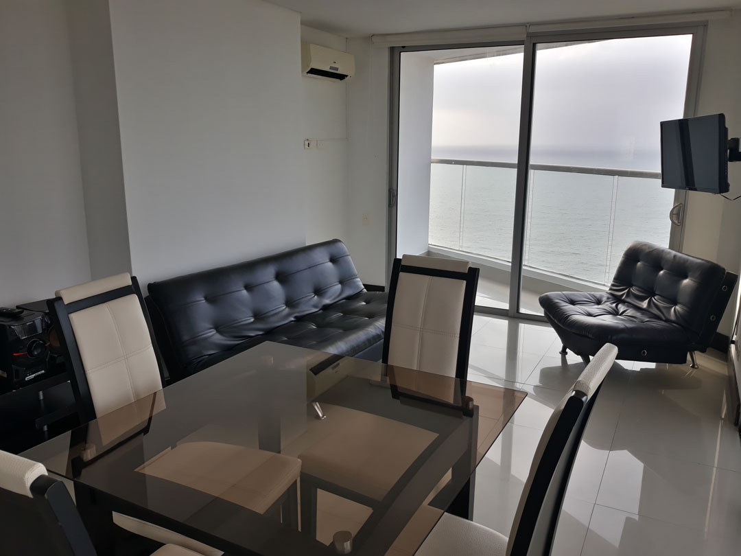 sala comedor de apartamento con mobiliario en cuero y vidrio, un ventanal piso techo da salida a un balcón con vista al mar