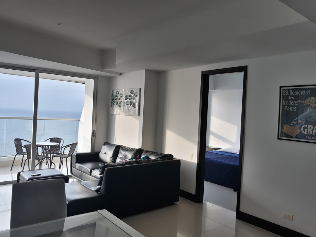 vista general de apartamento su sala está amoblada con sofá de cuero, habitación y balcón amoblado que tiene vista al mar