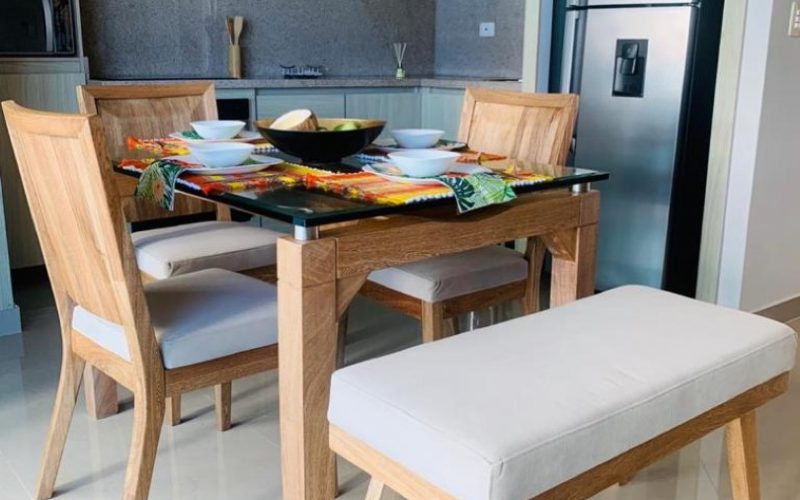 detalle de mesa comedor en madera y vidrio con 3 sillas y una butaca; al fondo acompaña una cocina abierta de estilo moderno