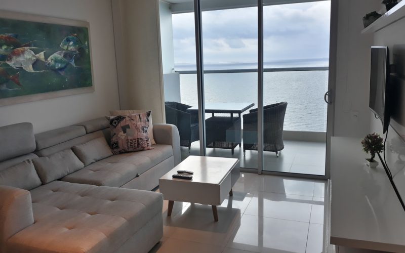 sala con sofa en ele gris y televisor en la pared al fondo balcon con muebles exteriores y vista a playa de bocagrande
