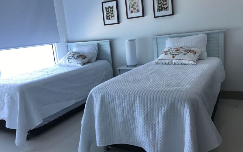 habitacion con dos cama sencillas en madera color blanco y una mesa de noche con lampara color blanco