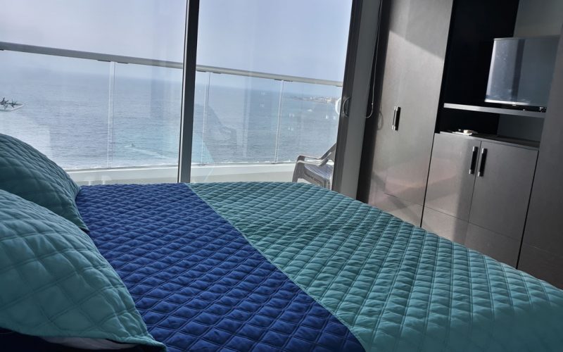 habitación con cama doble, armario y tv anidado. un ventanal de piso a techo da salida a balcón con vista al mar