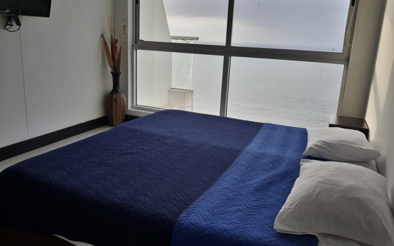 habitación con cama doble, nochero y decoración simple. un ventanal piso a techo da vista al mar