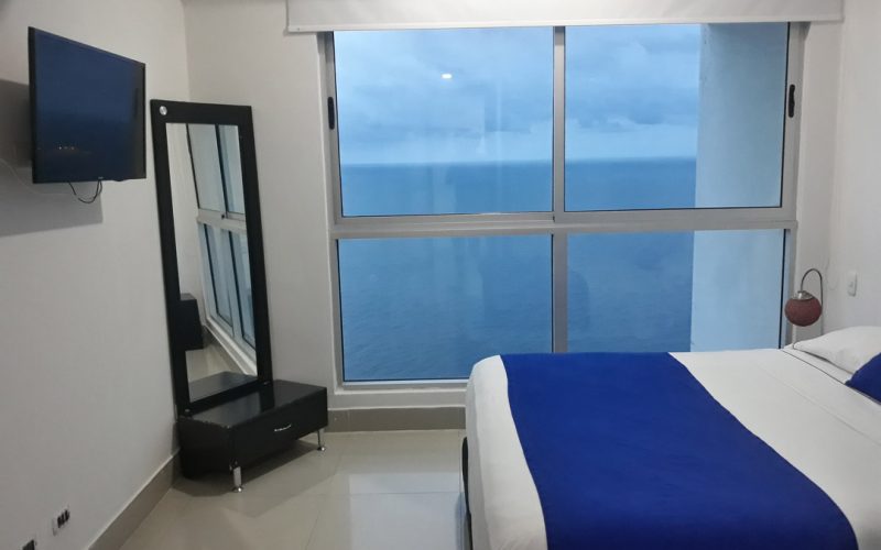 habitacion con cama doble y mesas de noche en madera, aire acondicionado, televisor y ventanal con vista al mar